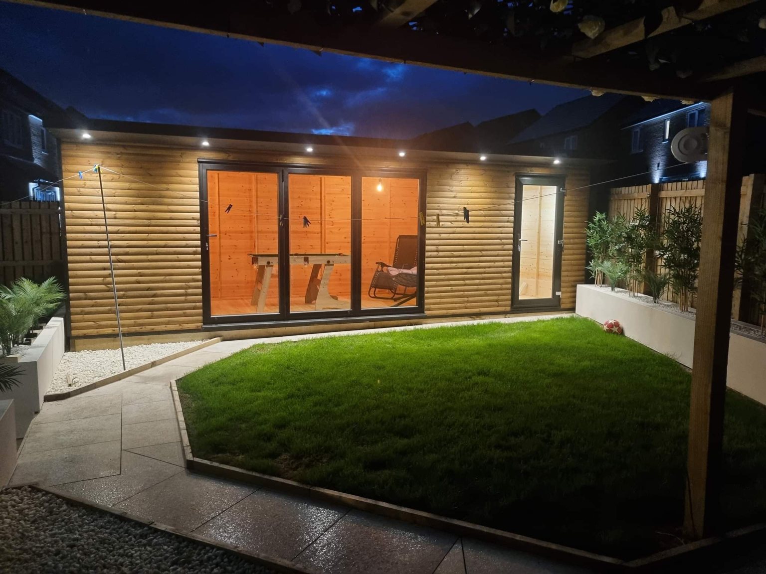 A garden room at dusk lit by a row of soffit downlights, with lighting along the fence and planters