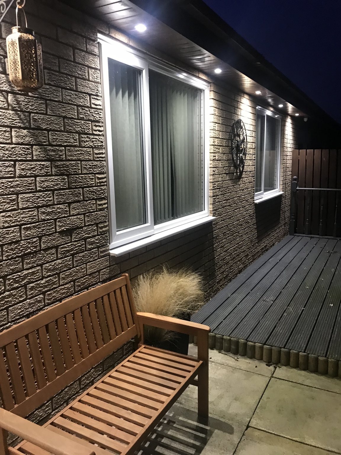 Recessed soffit downlights washing light down a brick bungalow wall at night, above a patio bench and decking