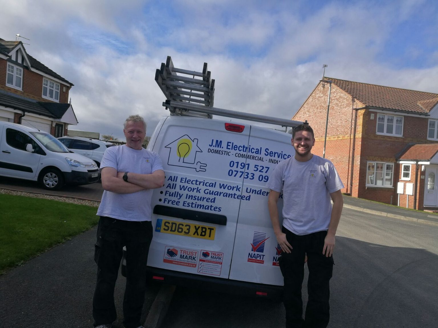 Two of the J M Electrical Services team in branded T-shirts standing either side of the liveried company van
